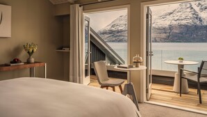 A bedroom with a large bed, a small table with a chair, a view of a mountain and lake through open doors. - Rosewood Matakauri (Closeburn)