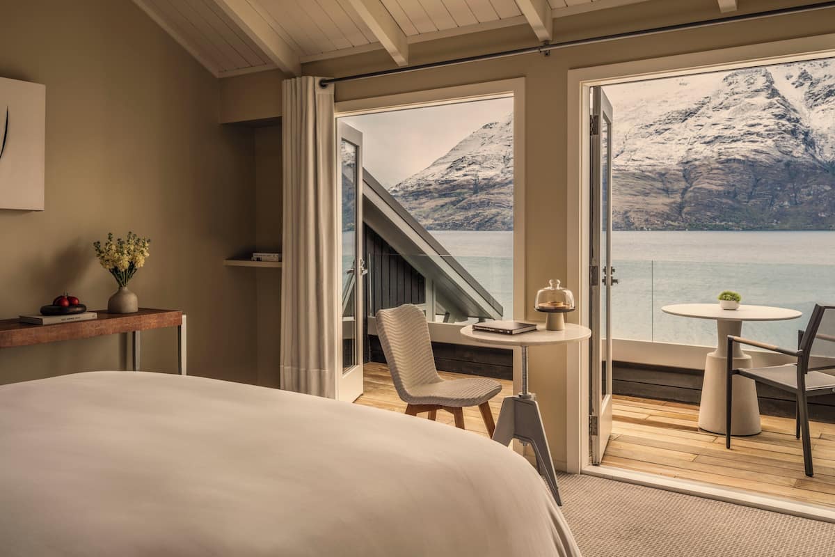 A bedroom with a large bed, a small table with a chair, a view of a mountain and lake through open doors.
