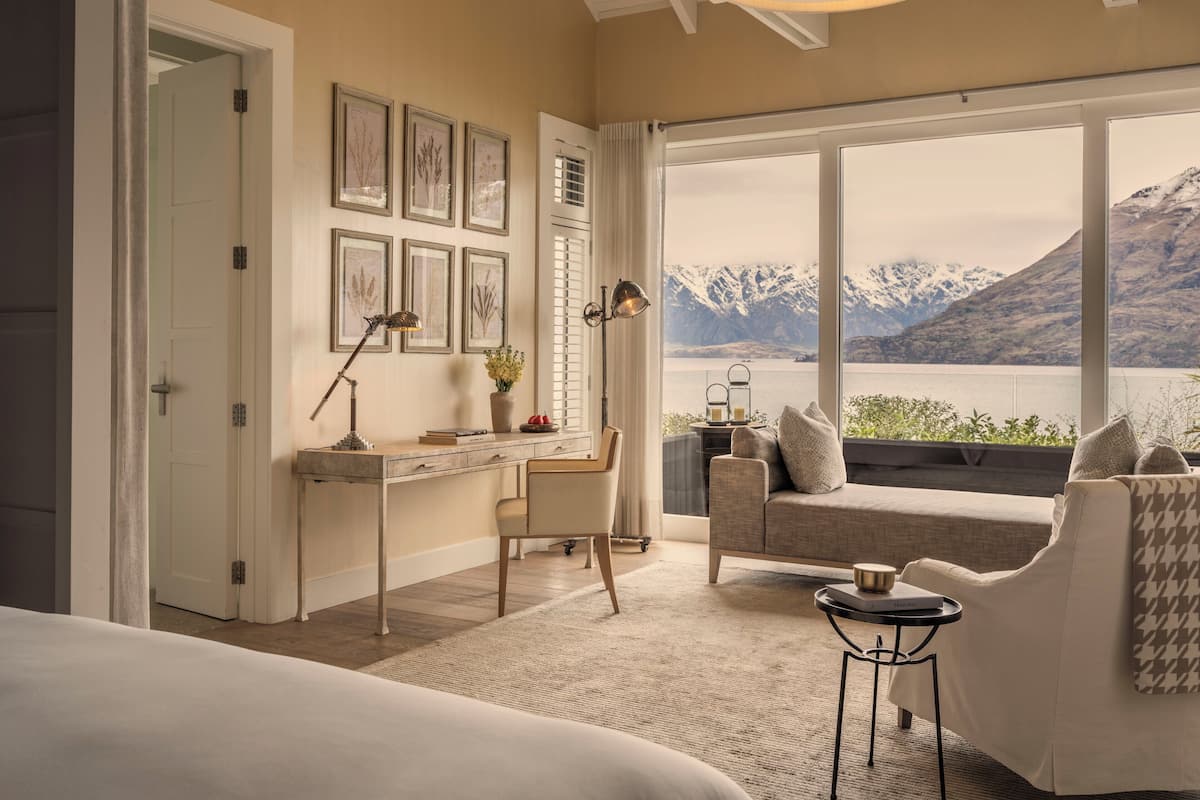 A bedroom with a large window, a desk, a chair, a bed, and a view of mountains and a lake.