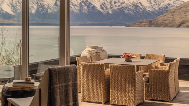 A cozy outdoor seating area with wicker furniture, a table with fruit, and a view of snow-capped mountains and a lake. - Rosewood Matakauri (Closeburn)