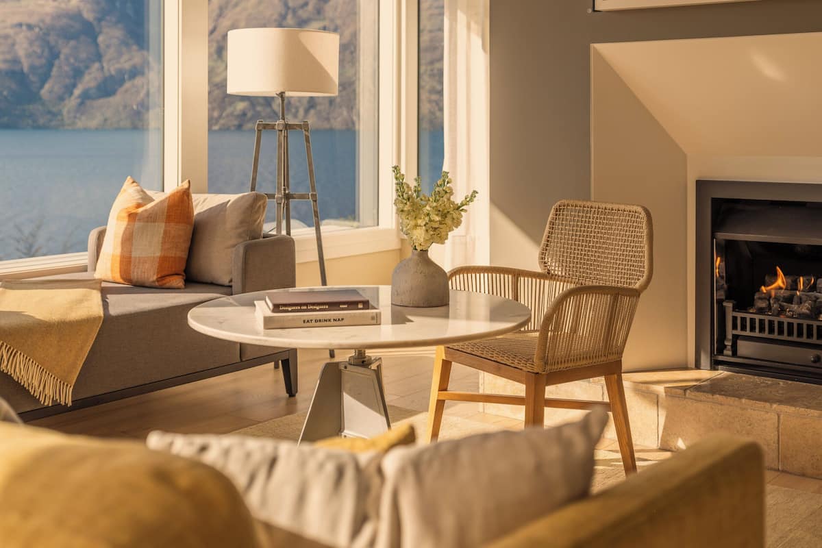 A modern living room with a fireplace, large windows with a mountain view, a cozy seating area, and a small table with books and a vase.