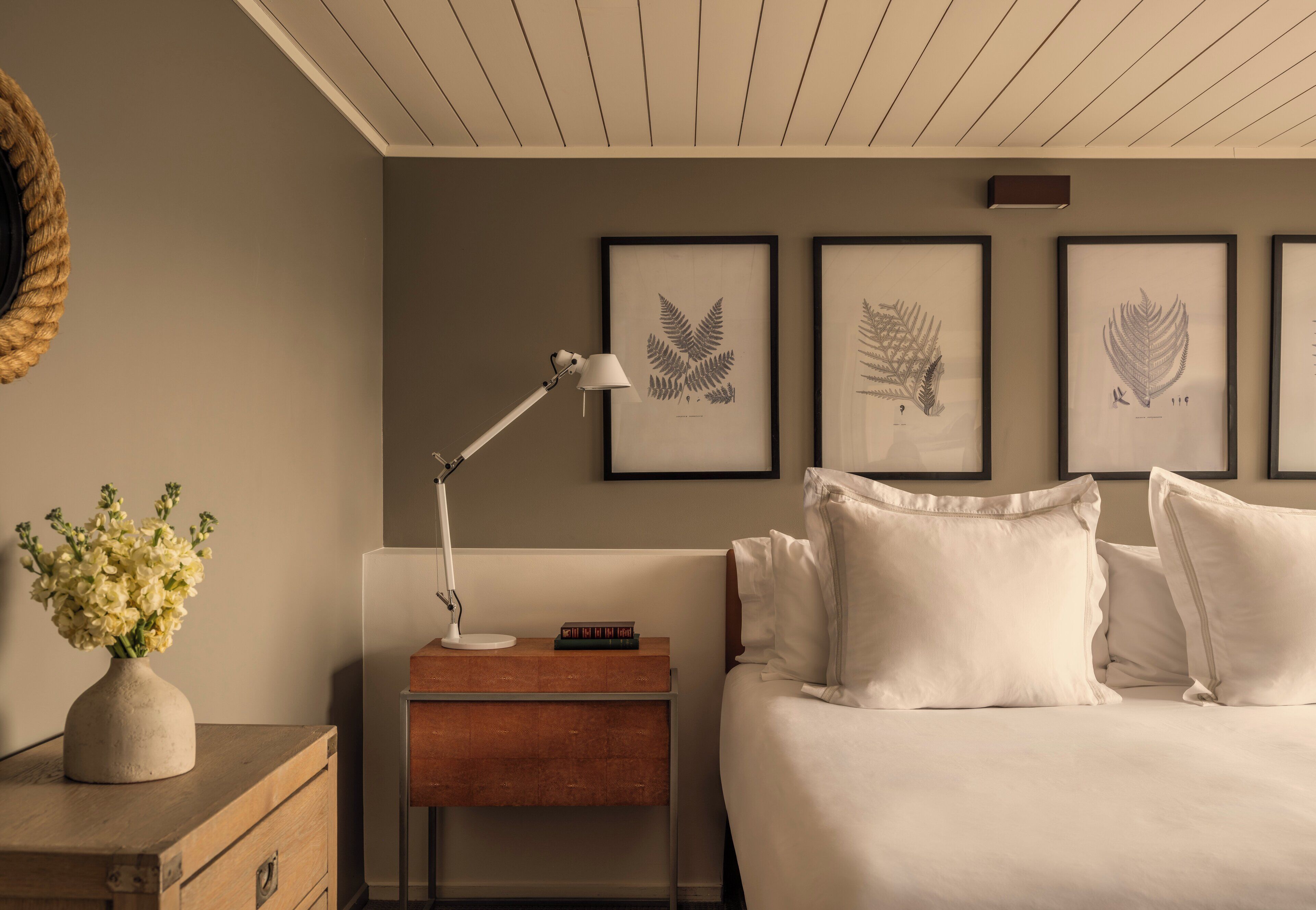 A bedroom with a bed, bedside table, lamp, framed botanical prints, and a decorative basket.