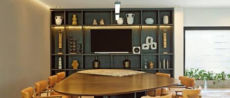 A modern dining area with a large round table surrounded by wooden chairs, a built-in TV, and decorative shelves with various items.
