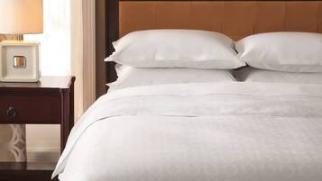 Una cama bien hecha con sábanas y almohadas blancas, una mesita de noche con una lámpara, y una pared con papel tapiz estampado.