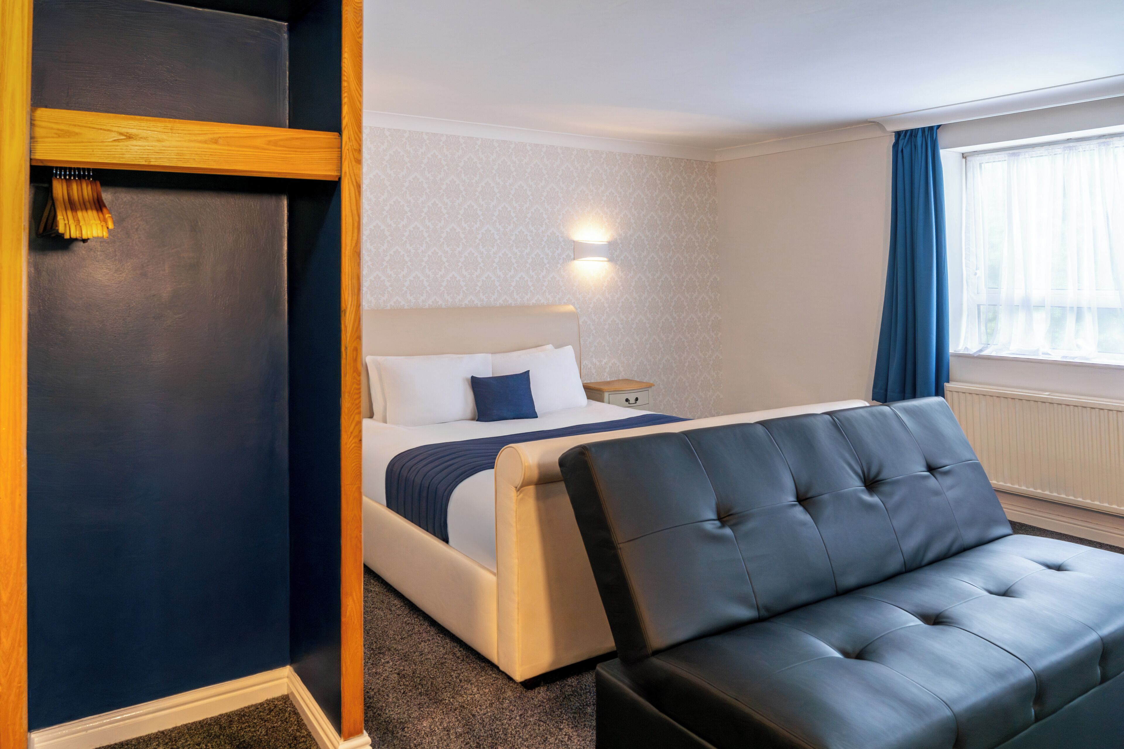 Habitación de hotel con cama, mesita de noche, lámpara, ventana con cortinas y un área de descanso con una silla.