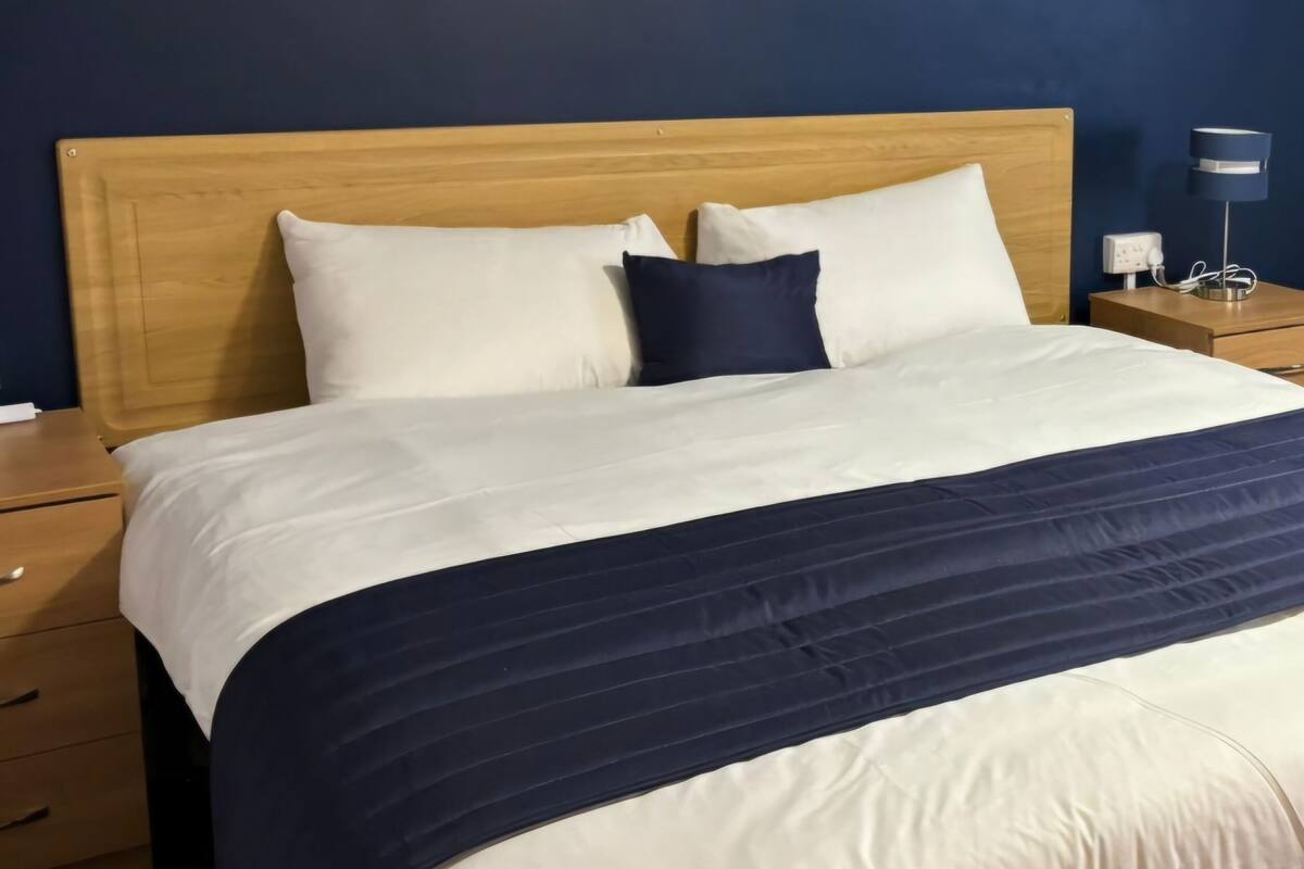 A neatly made bed with a wooden headboard, two white pillows, and a navy blue accent pillow. There is a bedside table with a lamp and a power outlet.