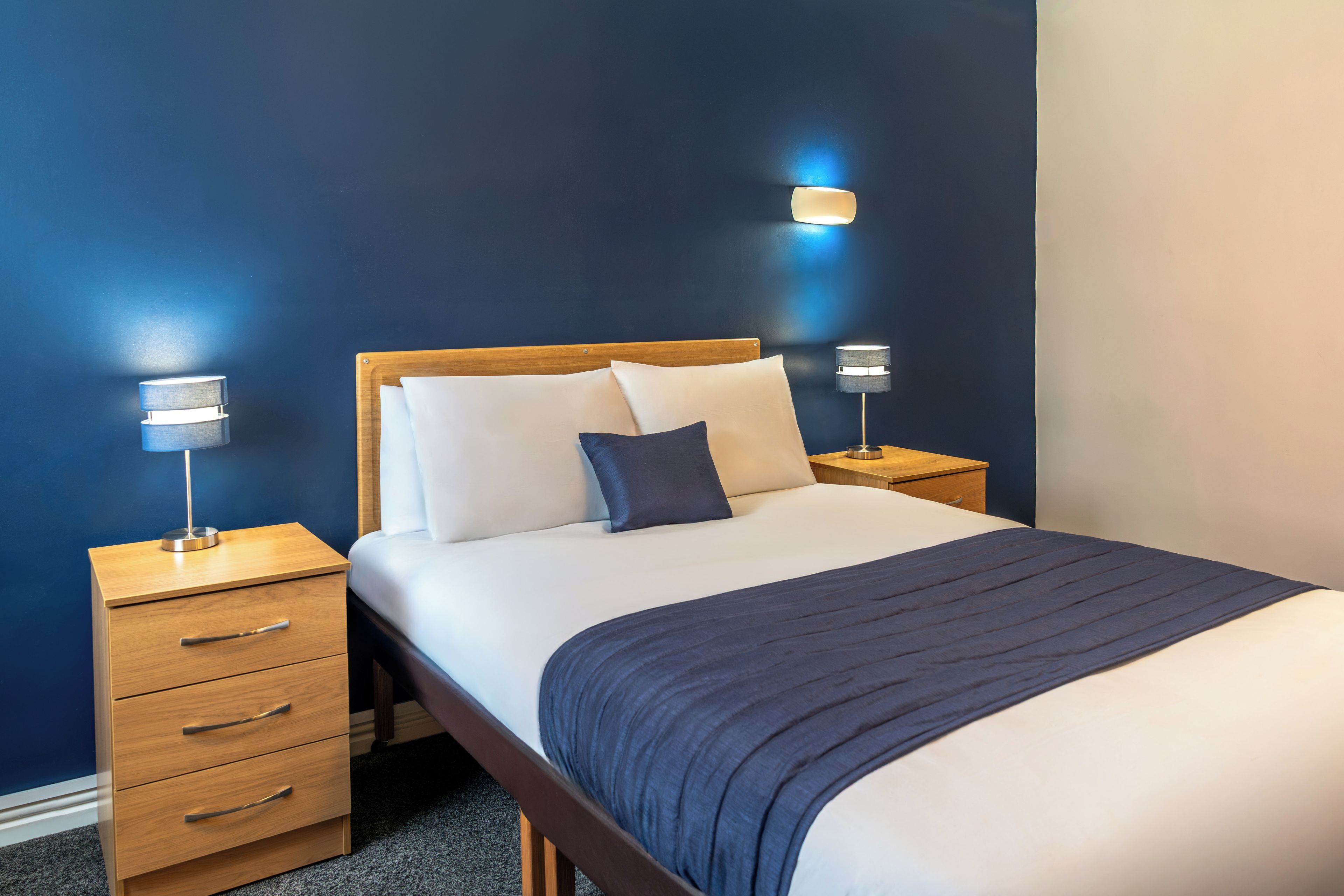 Ein ordentlich bezogenes Bett mit einem hölzernen Kopfteil, zwei Nachttische mit Lampen und eine dunkelblaue Wand.