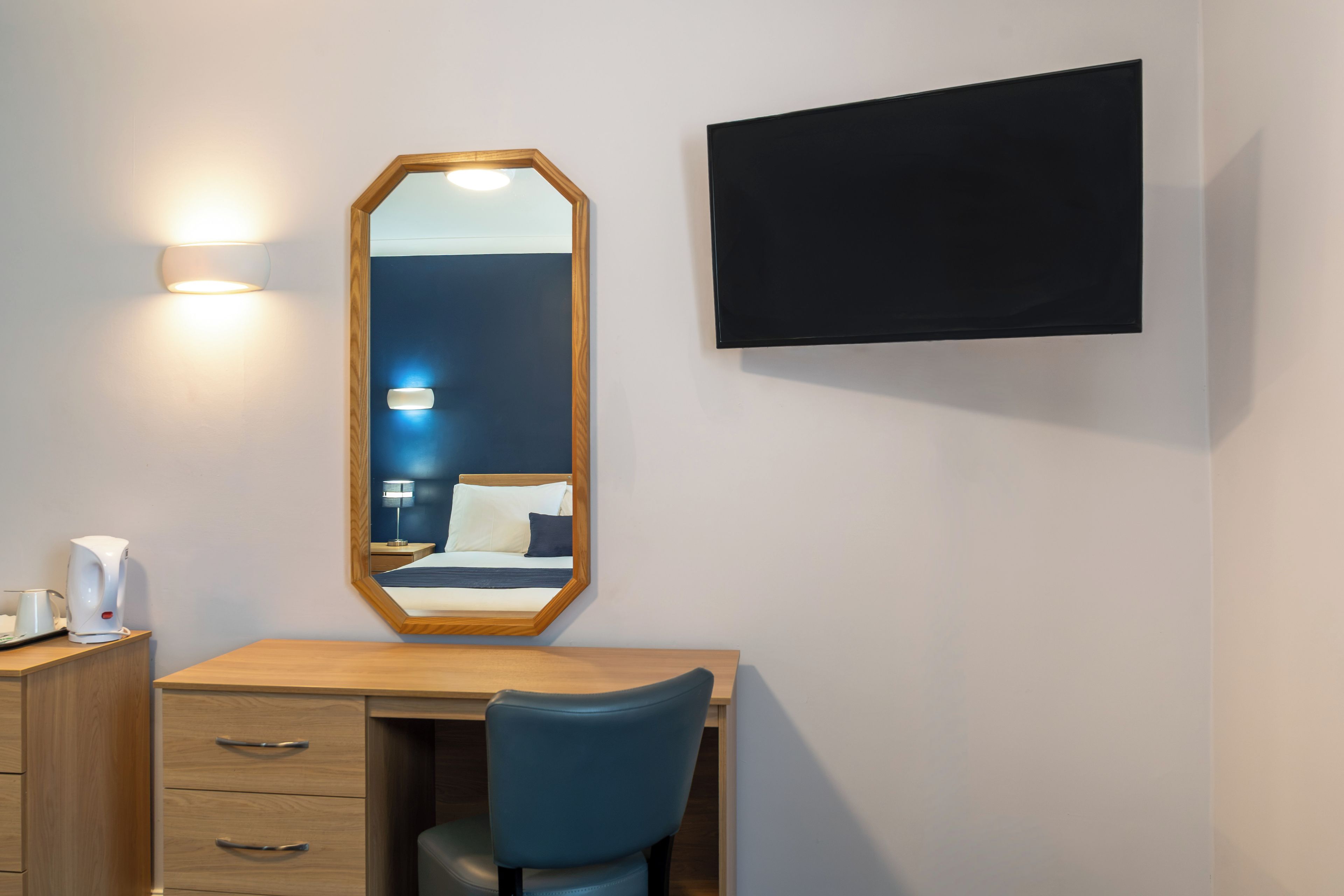 Ein Hotelzimmer mit Holztisch, einem Sessel, einem großen Spiegel, einer Nachttischlampe, einem Flachbildfernseher und einem Nachttisch mit Lampe.