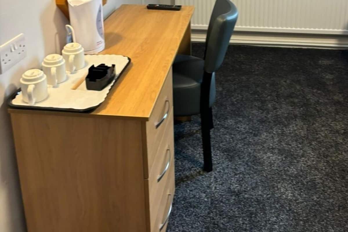A hotel room with a wooden desk, a chair, a flat-screen TV mounted on the wall, a radiator, and a tray with cups and a phone on the desk.