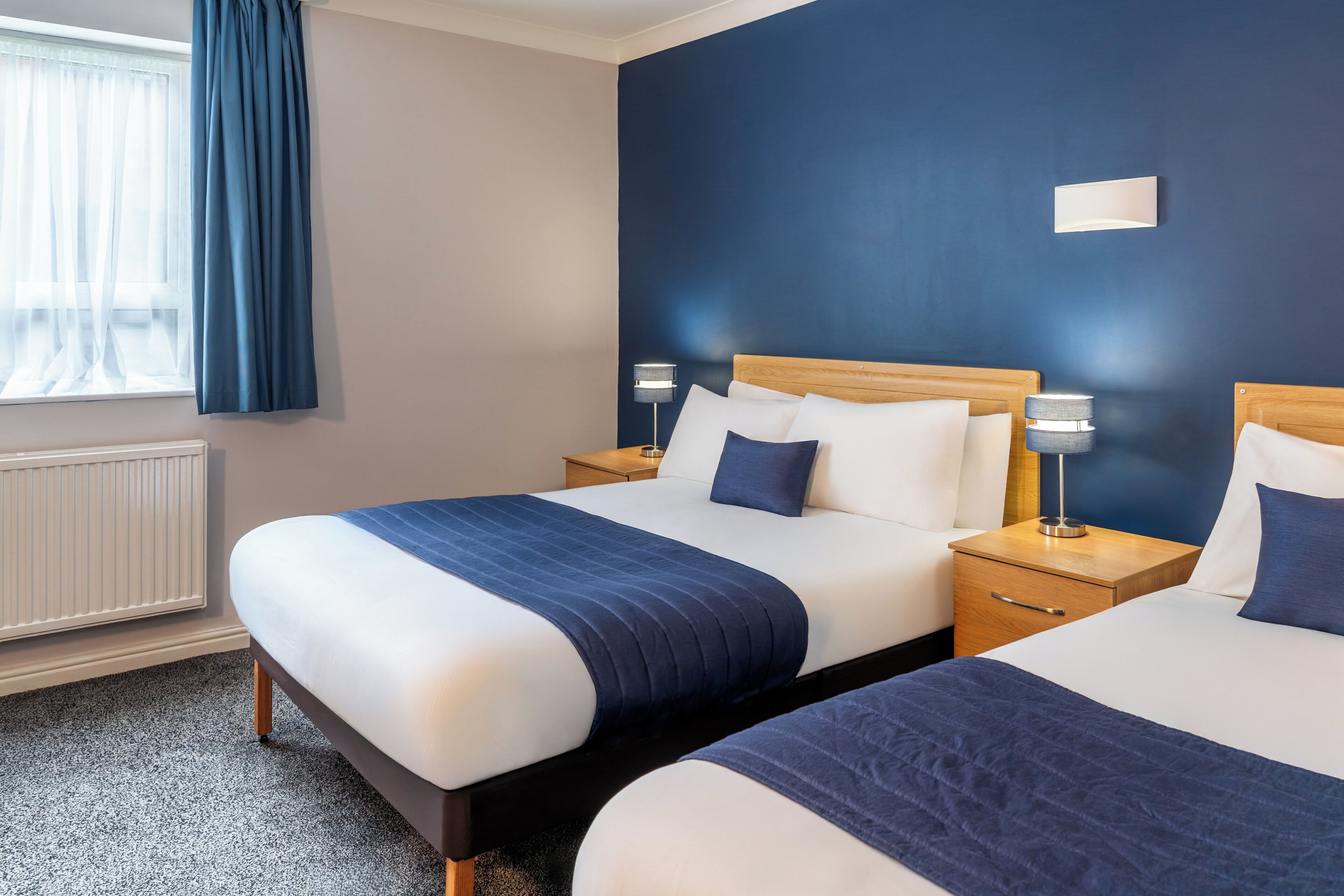 Habitación de hotel con dos camas, una mesita de noche con lámpara, una ventana con cortinas y un radiador.