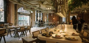 A restaurant with a long counter, multiple dining tables, and chairs. There are plates of food on the counter, and a person is standing near the counter.
