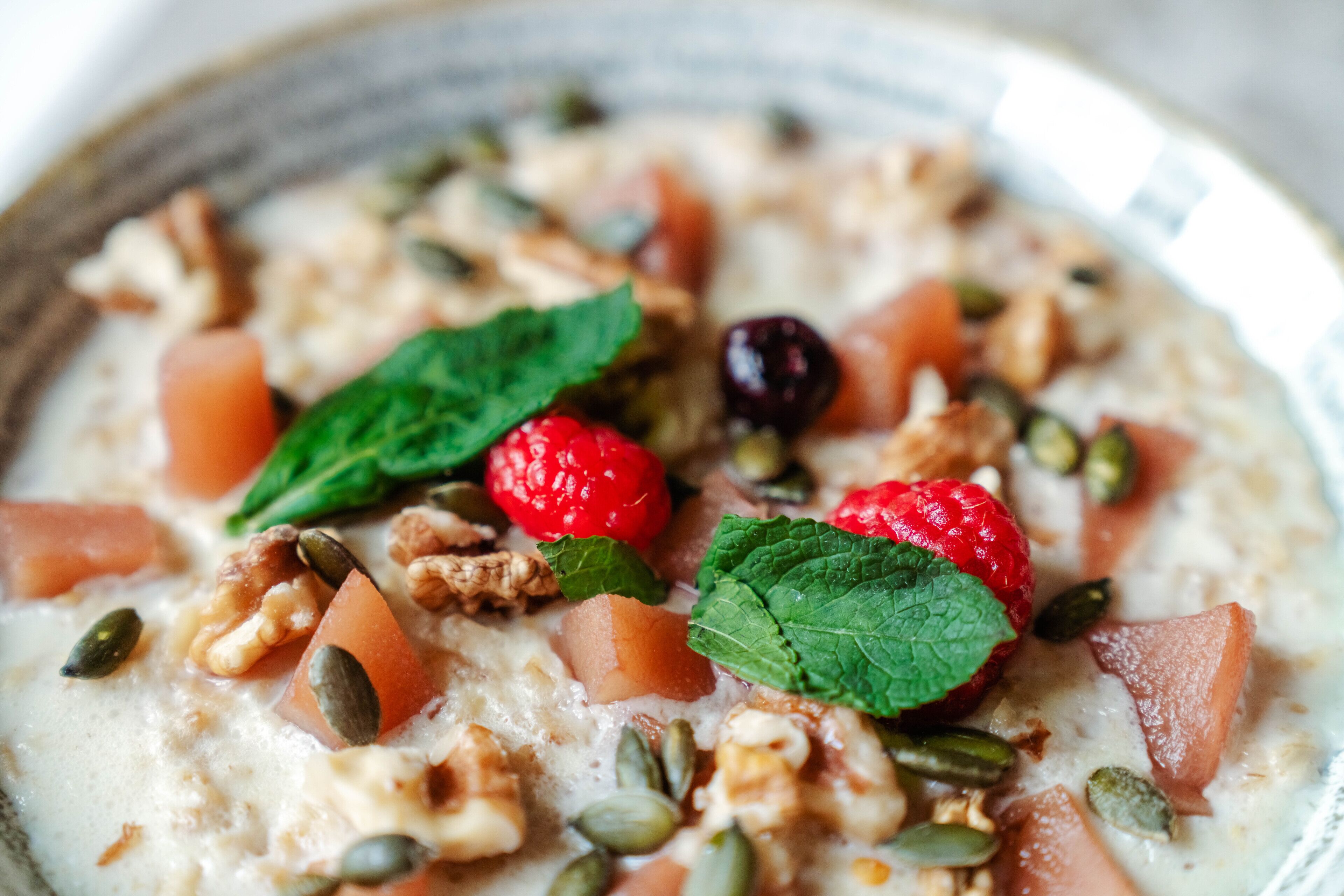 A bowl of oatmeal with raspberries, walnuts, pumpkin seeds, and mint leaves.