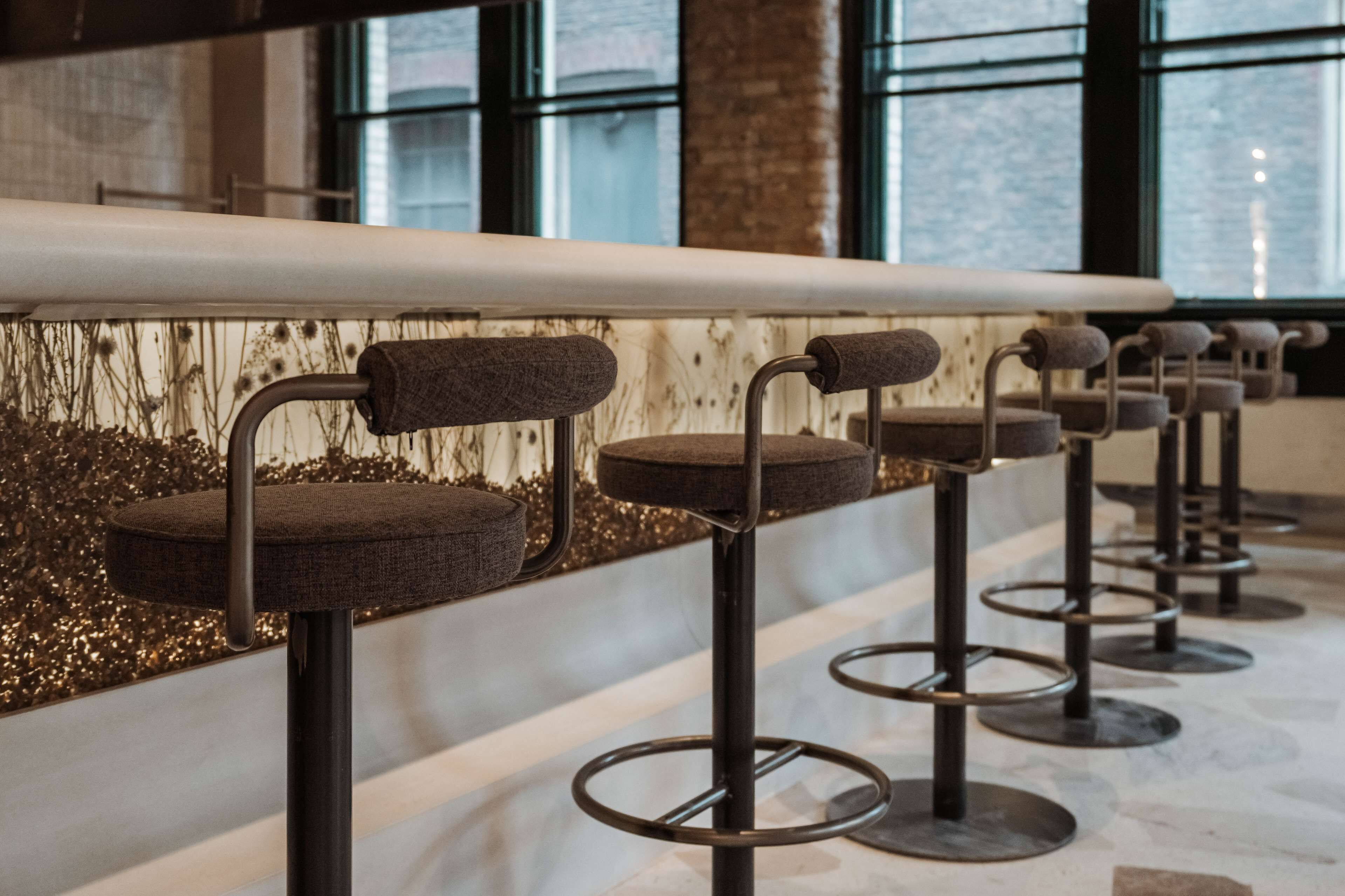 Fila de taburetes con asientos acolchados y patas metálicas, ubicados en un espacio interior moderno con suelo de mármol y una pared decorativa con detalles dorados.
