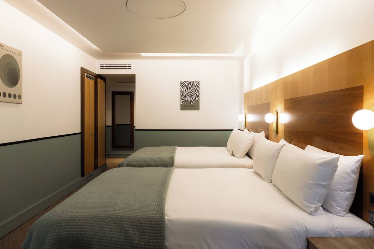 A hotel room with two beds, a wooden headboard, a wall-mounted artwork, and a door leading to another room.