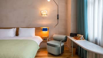 A hotel room with a bed, a chair, a table, a wall-mounted lamp, and a window with curtains.