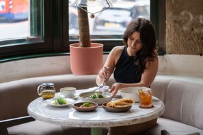A woman dining at a round table with various dishes, a lamp, and a view of the street through a window.