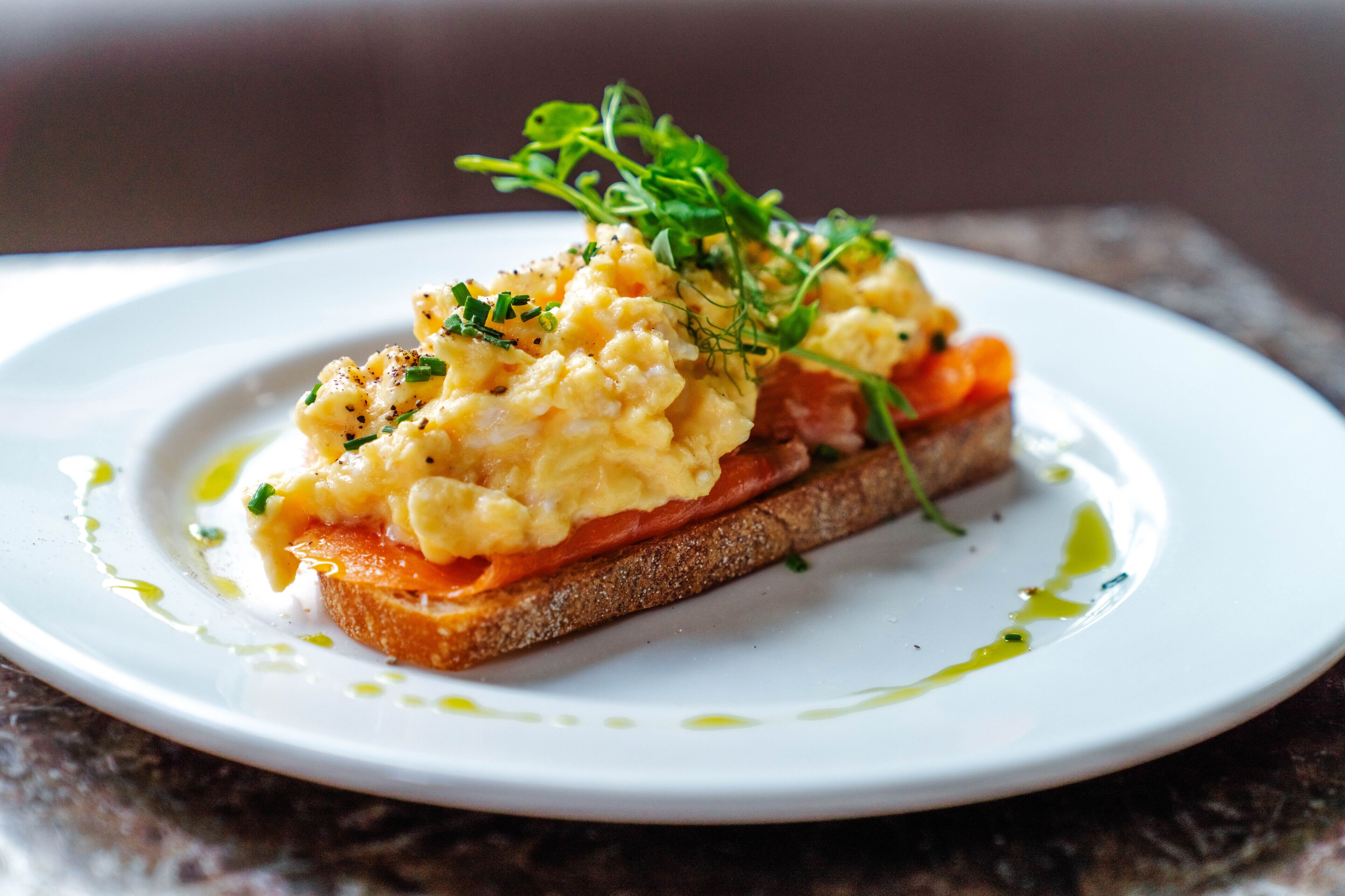 A plate with scrambled eggs, smoked salmon, and a garnish on a slice of bread.