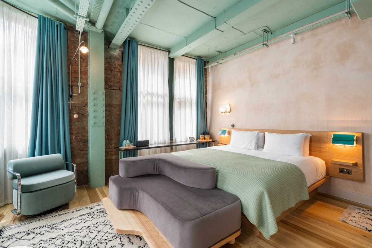 A modern bedroom with a large bed, a sofa, and a chair. There is a window with curtains, a bedside table, and a wall-mounted light.