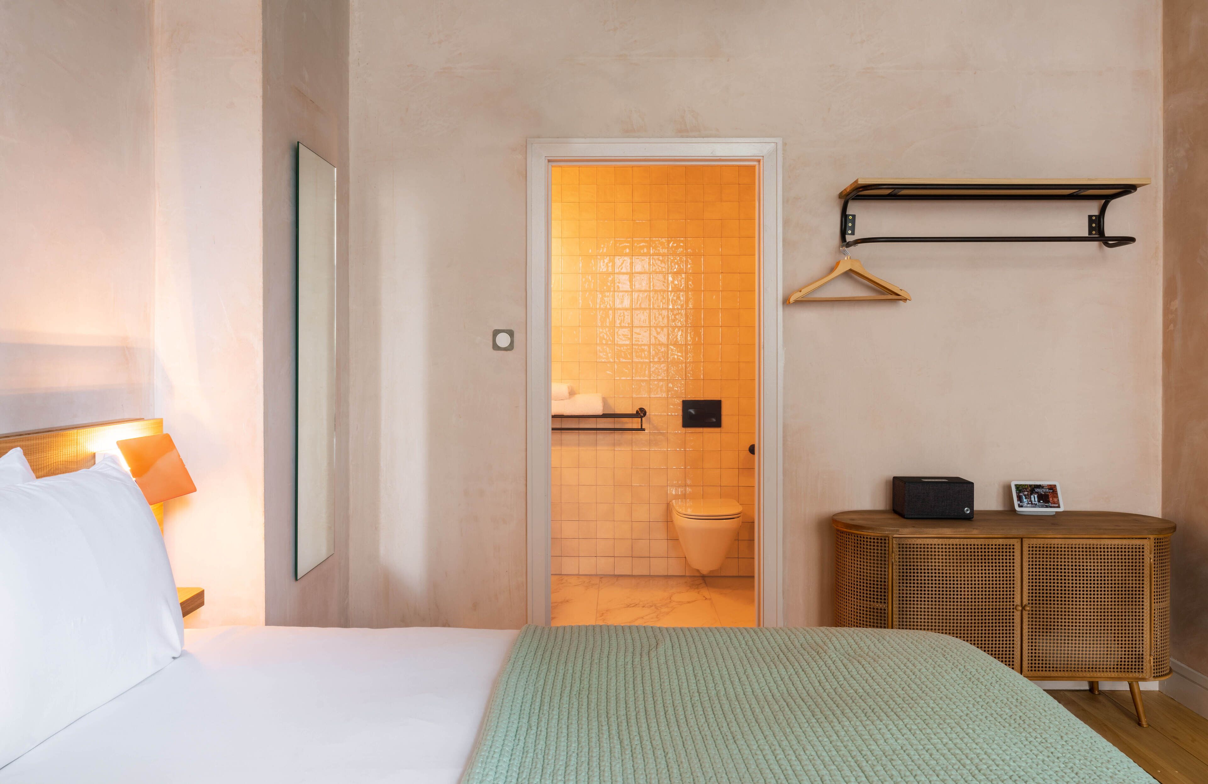 A hotel room with a bed, a bedside lamp, a mirror, a wooden side table, a hanging coat rack, and a view into the bathroom.