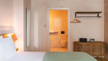 A hotel room with a bed, a bedside lamp, a mirror, a wooden side table, a hanging coat rack, and a view into the bathroom.