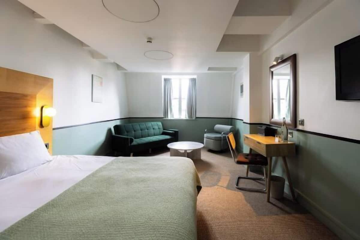 A hotel room with a bed, a green sofa, a small round table, a desk, and a mirror.