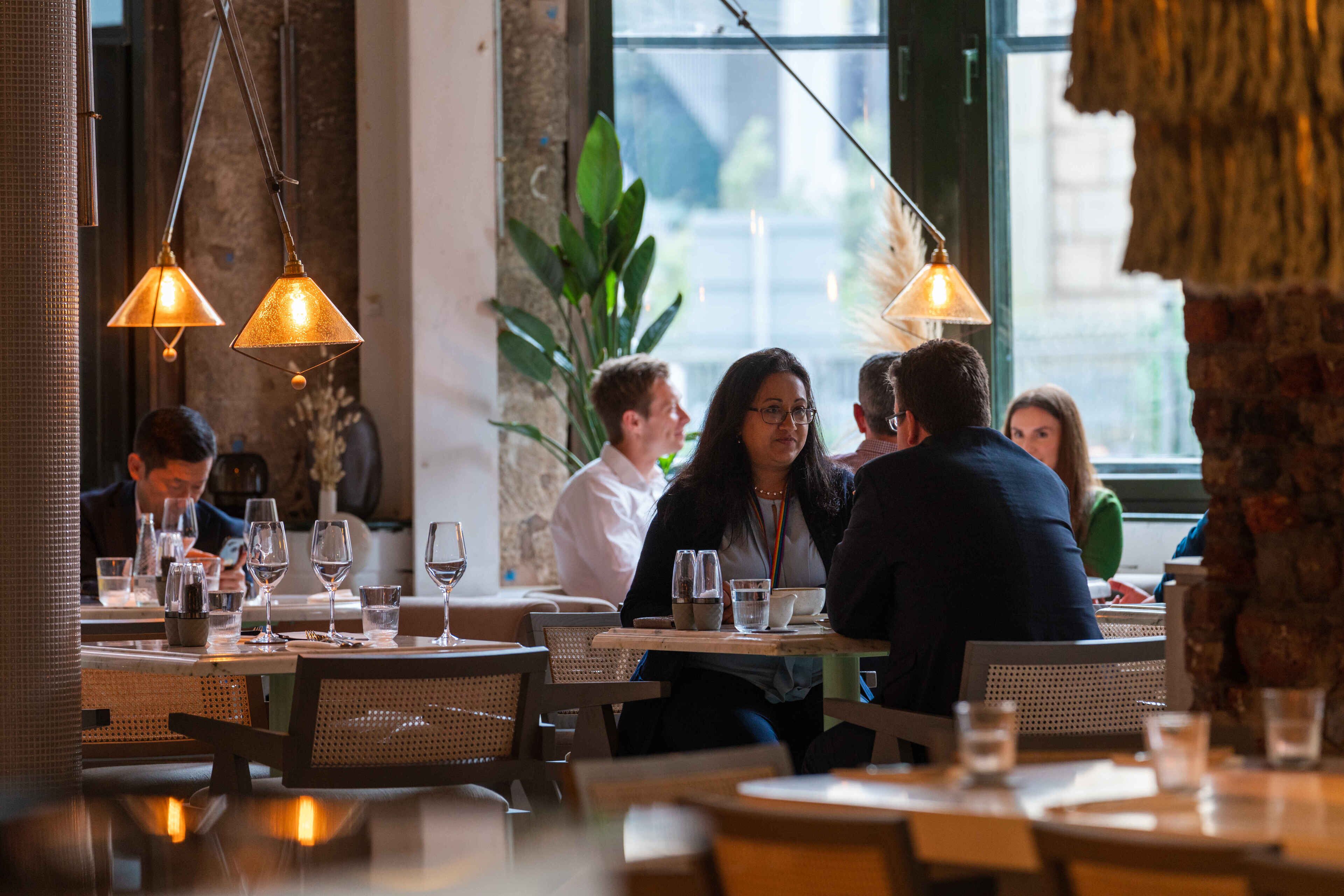 L’intérieur d’un restaurant est agrémenté de tables rondes, de luminaires suspendus et offre une vue sur l’extérieur grâce à de grandes fenêtres.