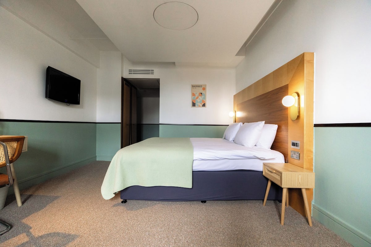 A hotel room with a bed, a bedside table, a chair, a television, and a poster on the wall.