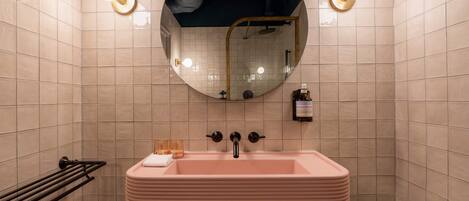 A bathroom with a pink sink, two wall-mounted lights, a round mirror, and a towel rack.