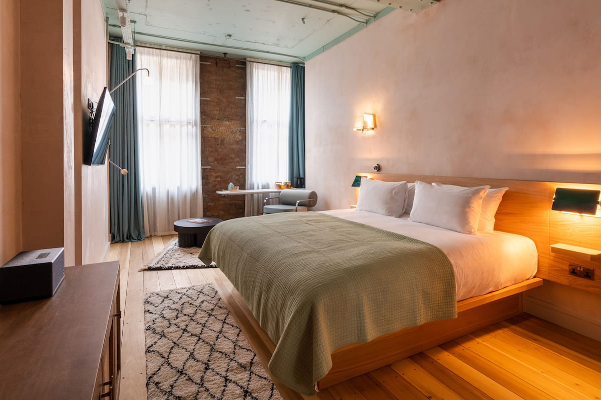 A modern bedroom with a wooden bed, a desk, and a chair. There is a window with curtains and a wall-mounted light.