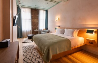 Un dormitorio moderno con una cama de madera, un escritorio y una silla. Se observa una ventana con cortinas y una lámpara fijada a la pared.