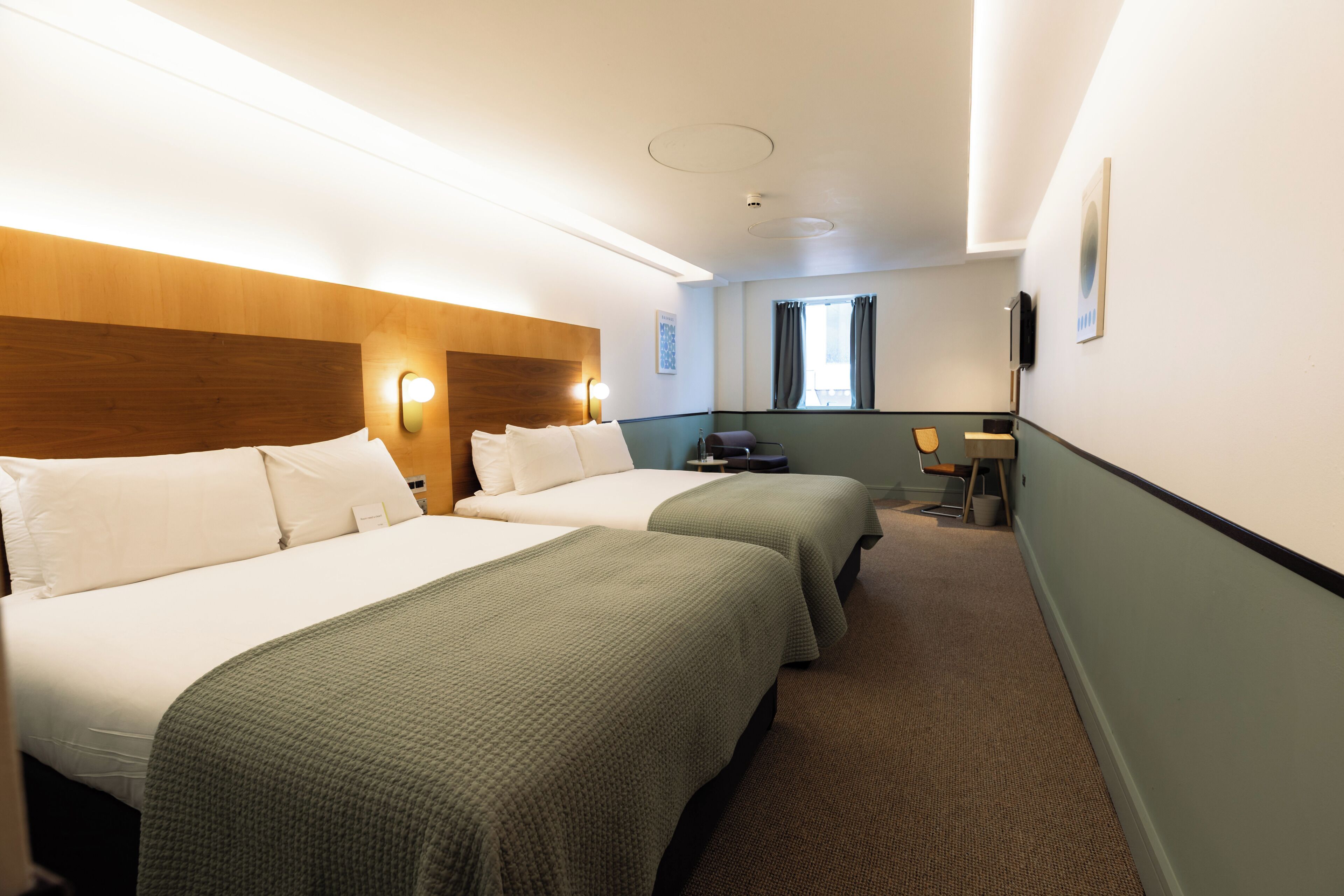 Habitación de hotel con dos camas, cabecera de madera, un escritorio pequeño y una ventana con cortinas.