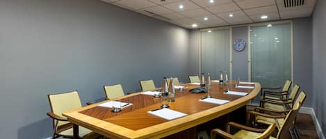 Un salón de conferencias con una amplia mesa de madera, sillas color beige, un reloj en la pared y una ventana con persianas.