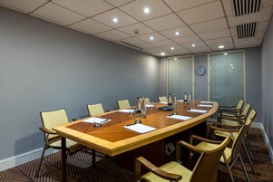 A conference room with a large wooden table, beige chairs, a clock on the wall, and a window with blinds.