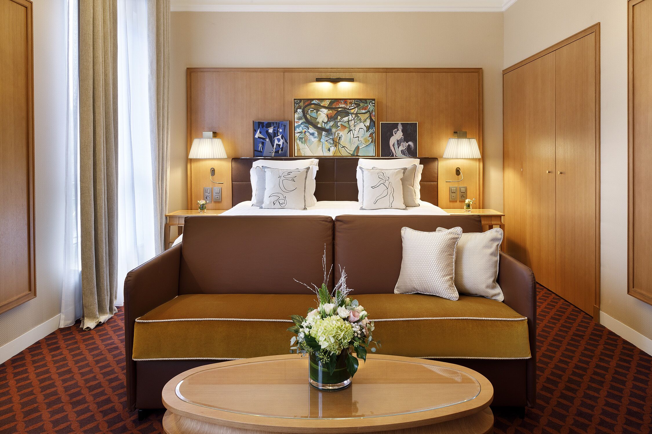 Une chambre d’hôtel avec un canapé marron, une table basse ronde agrémentée d’un arrangement floral, et des œuvres d’art accrochées au-dessus de la tête de lit.