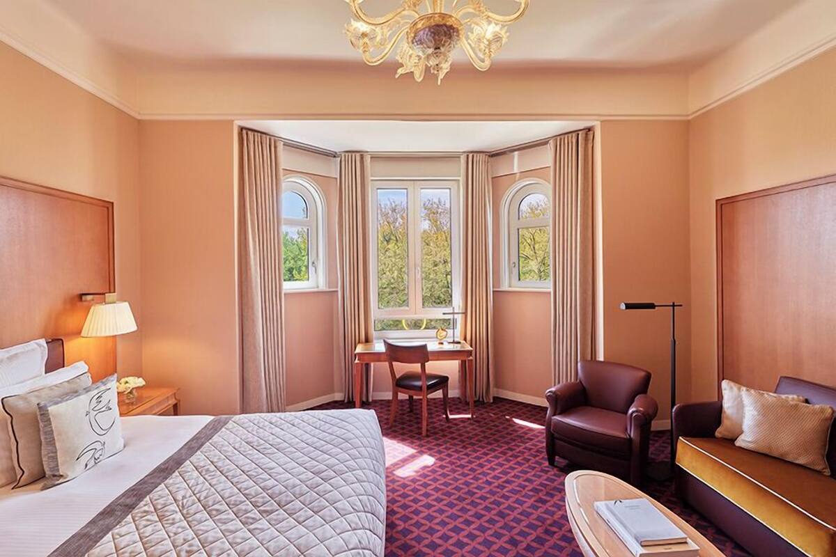 A hotel room with a large bed, a desk, a leather armchair, and a sofa. There are two windows with curtains, a chandelier, and a floor lamp.
