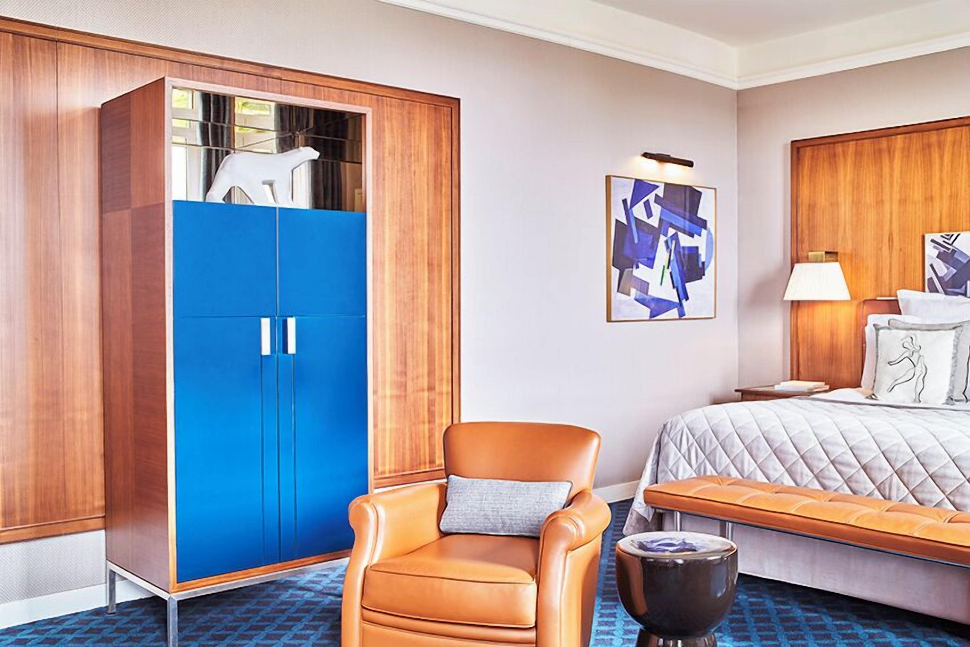A hotel room with a bed, a blue wardrobe, a leather armchair, a small round table, and a wall-mounted lamp.
