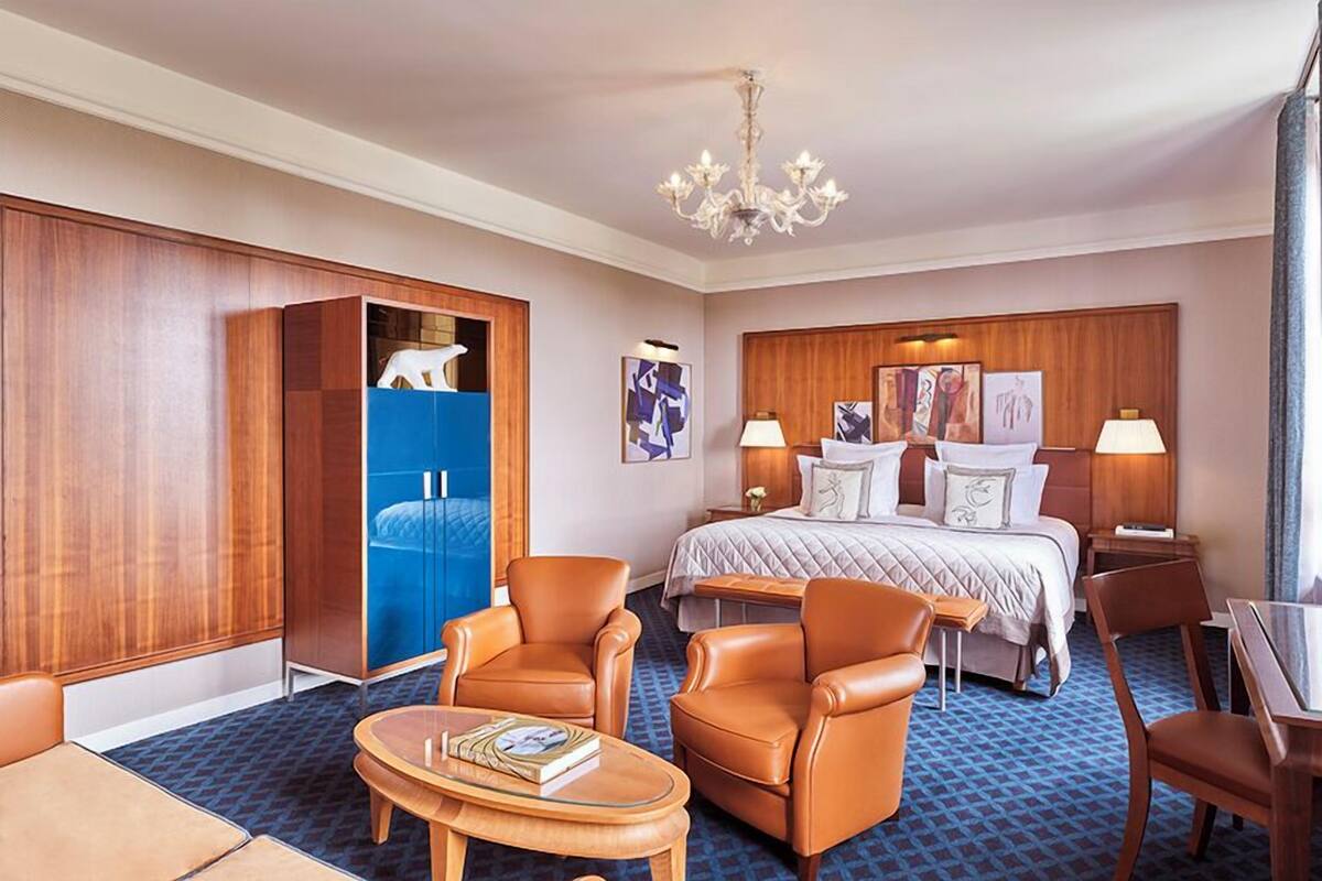 A hotel room with a bed, two armchairs, a small table, a wardrobe, and artwork on the walls.