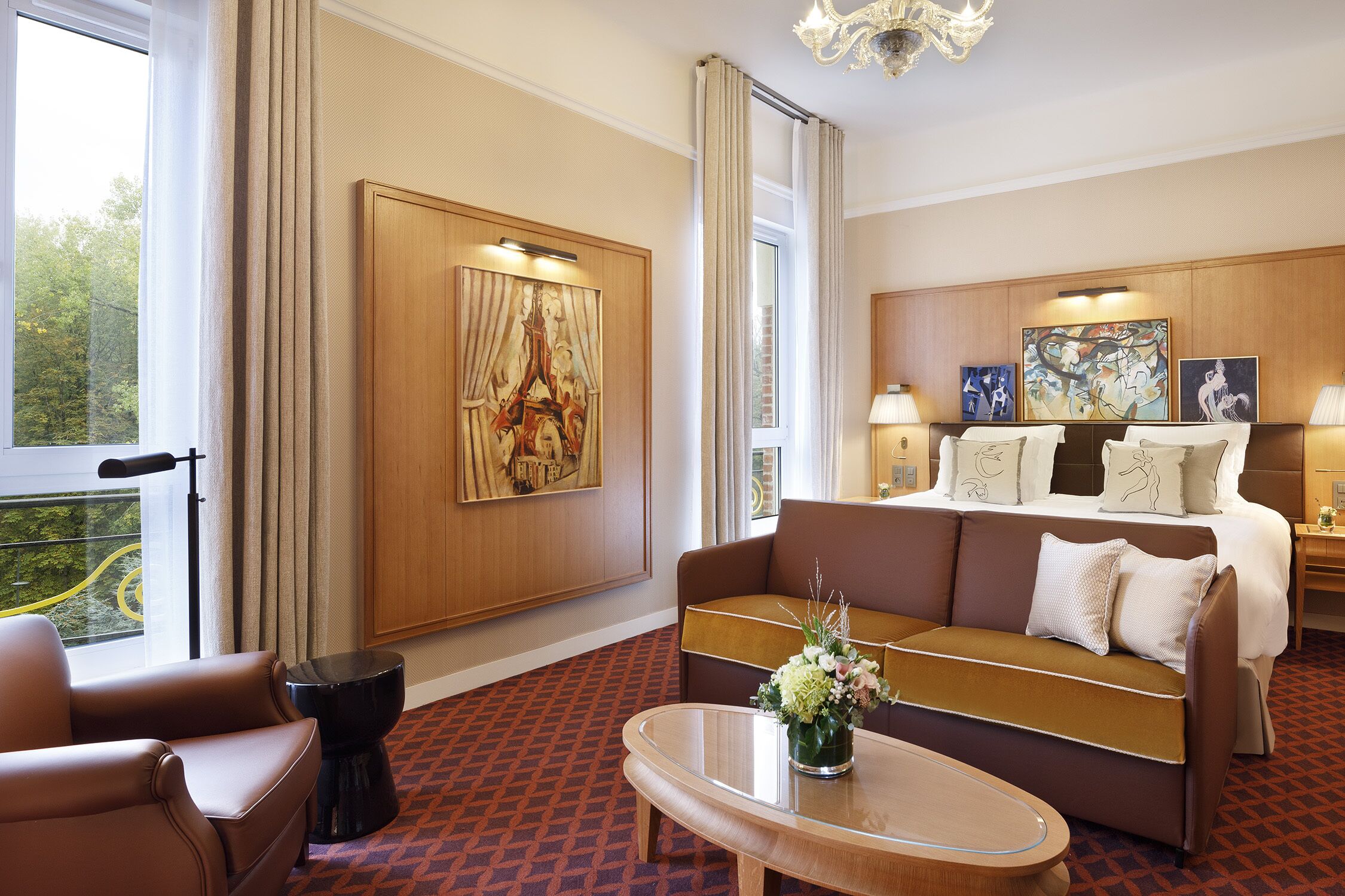A hotel room with a large bed, a sofa, a small table, and a window with a view of greenery.