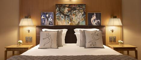 Habitación de hotel con una cama grande, dos mesitas de noche con lámparas, un cabecero de madera y tres obras de arte abstracto.