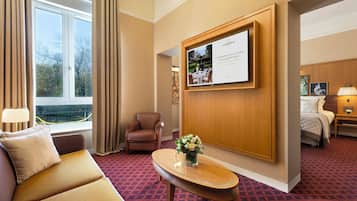 Une chambre d’hôtel avec un canapé, un fauteuil et une petite table sur laquelle est posé un vase de fleurs. On y trouve un lit avec une table de chevet et une lampe. Un grand tableau encadré orne le mur, et une fenêtre avec des rideaux est visible.