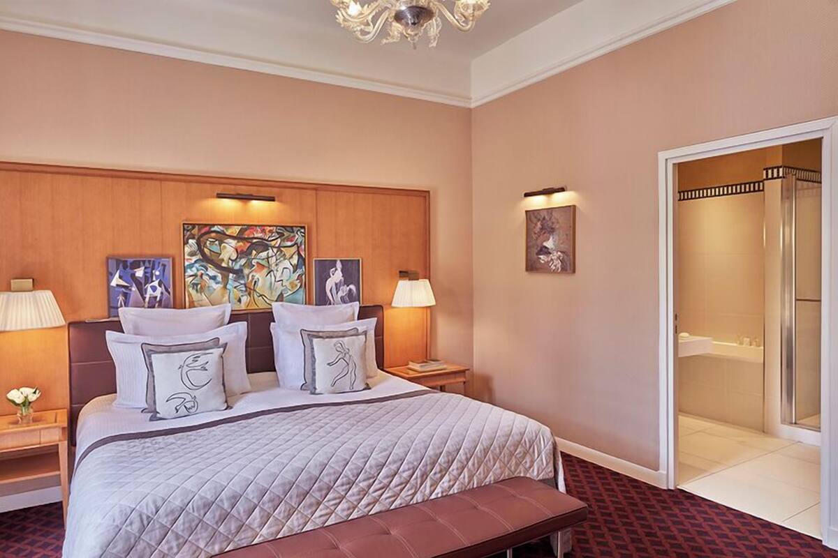 A hotel room with a large bed, two bedside lamps, a bench, a wooden headboard, and artwork on the walls.