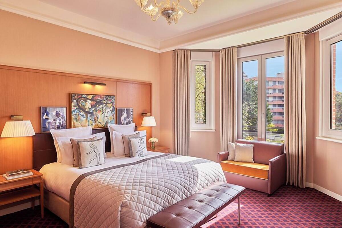 A hotel room with a large bed, a sofa, a bench, and a view of the outside.