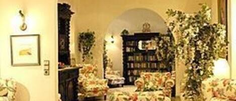 Ein gemütliches Wohnzimmer mit floralen Ohrensesseln, einem Bücherregal und einer großen Pflanze.