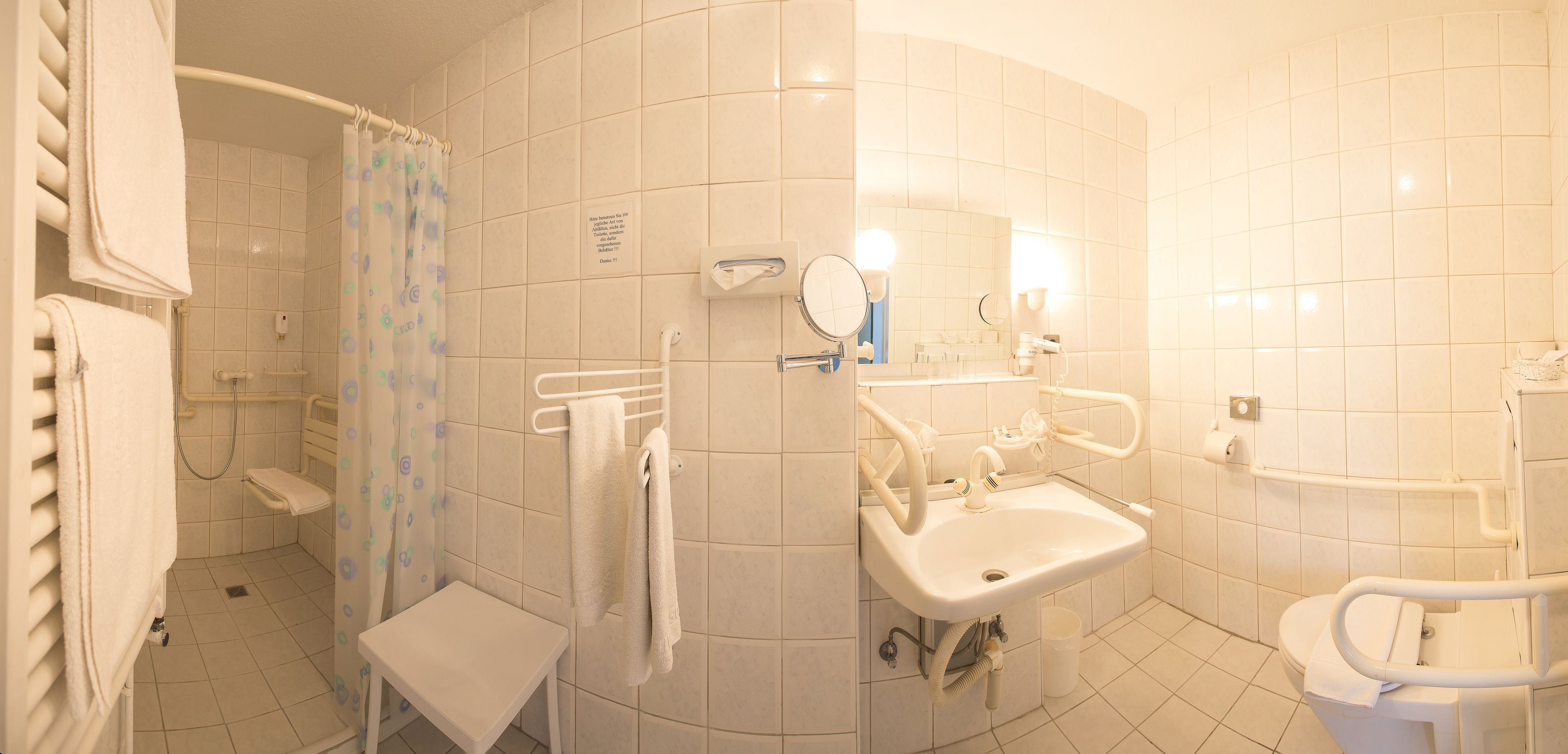 A bathroom with a shower, sink, and toilet. There is a towel rack and a bench.