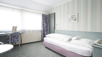 A hotel room with a bed, a desk, a chair, a radiator, and a window with curtains.
