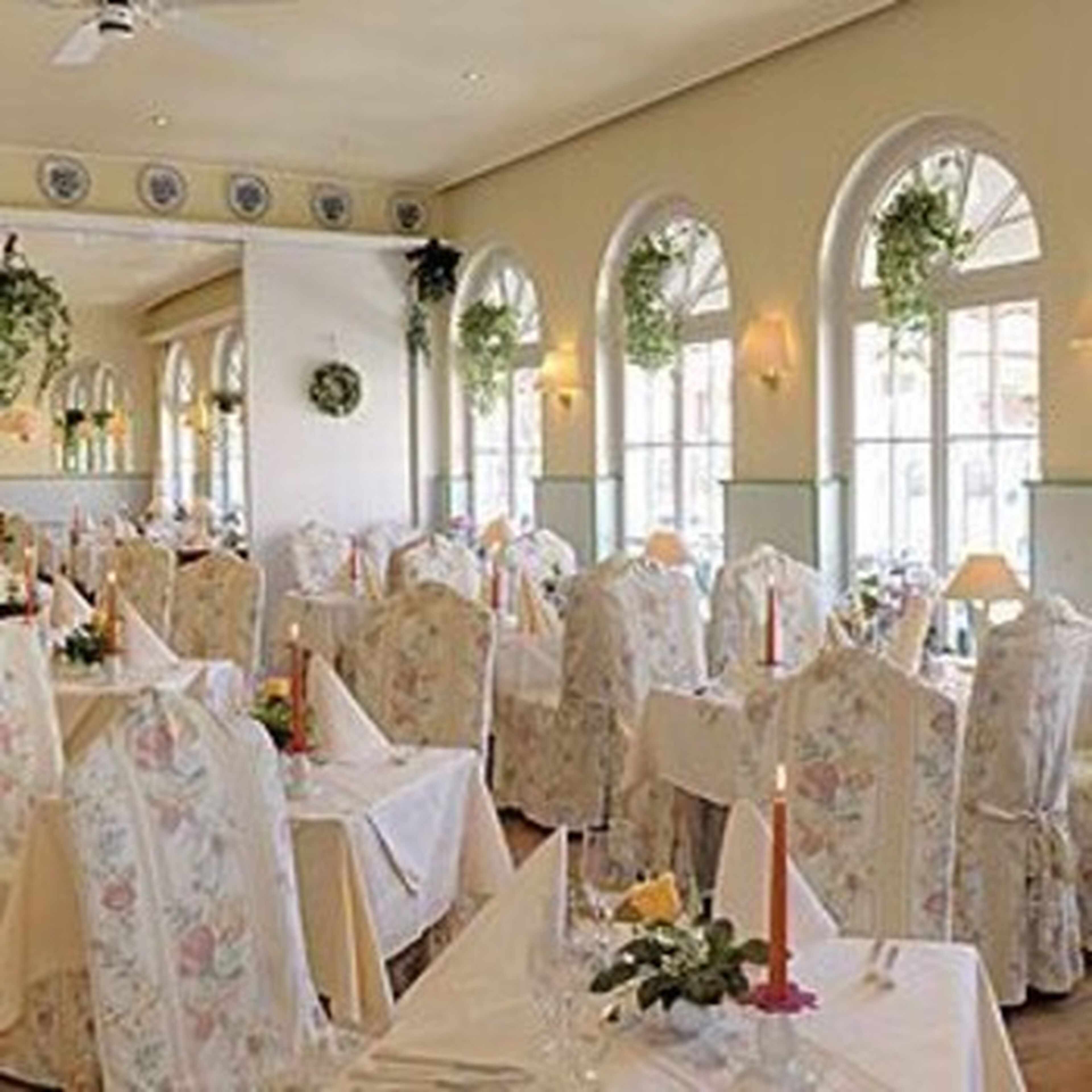 A banquet hall with round tables, floral chairs, and large arched windows.