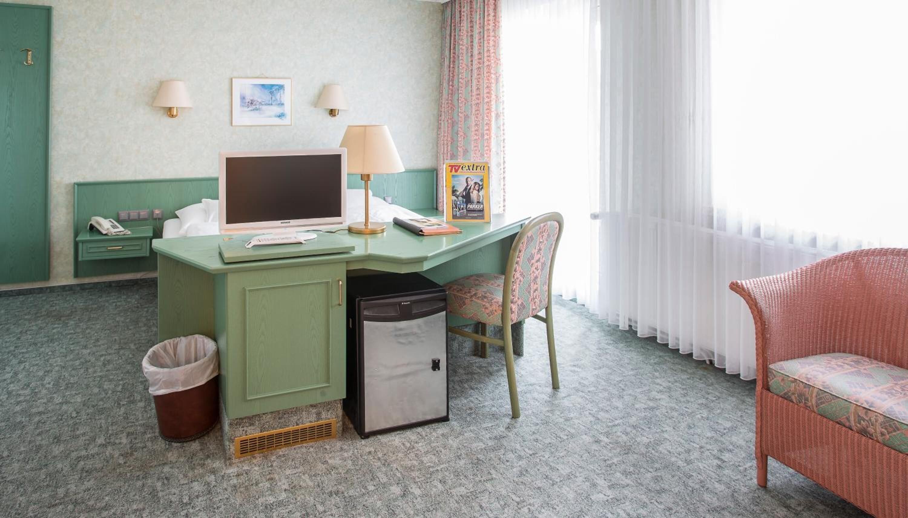 A hotel room with a green desk, a computer, a chair, a trash bin, and a wicker chair.