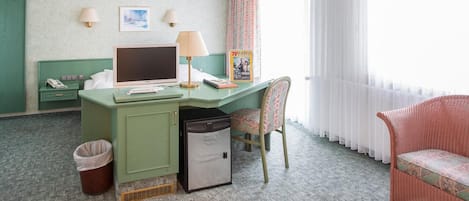A hotel room with a green desk, a computer, a chair, a trash bin, and a wicker chair.