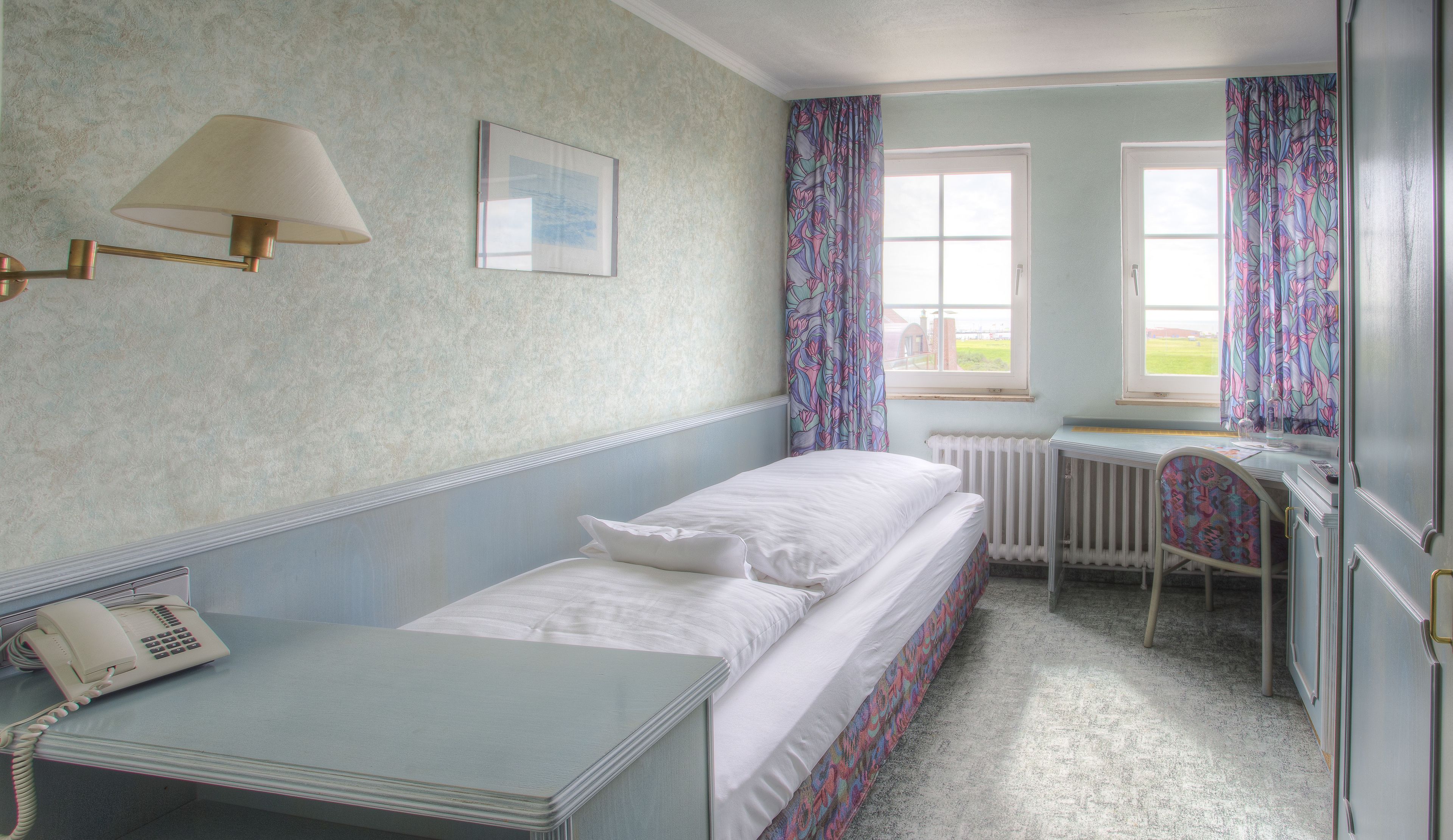 A single bed with white linens, a bedside table with a telephone, a radiator, and a window with floral curtains.