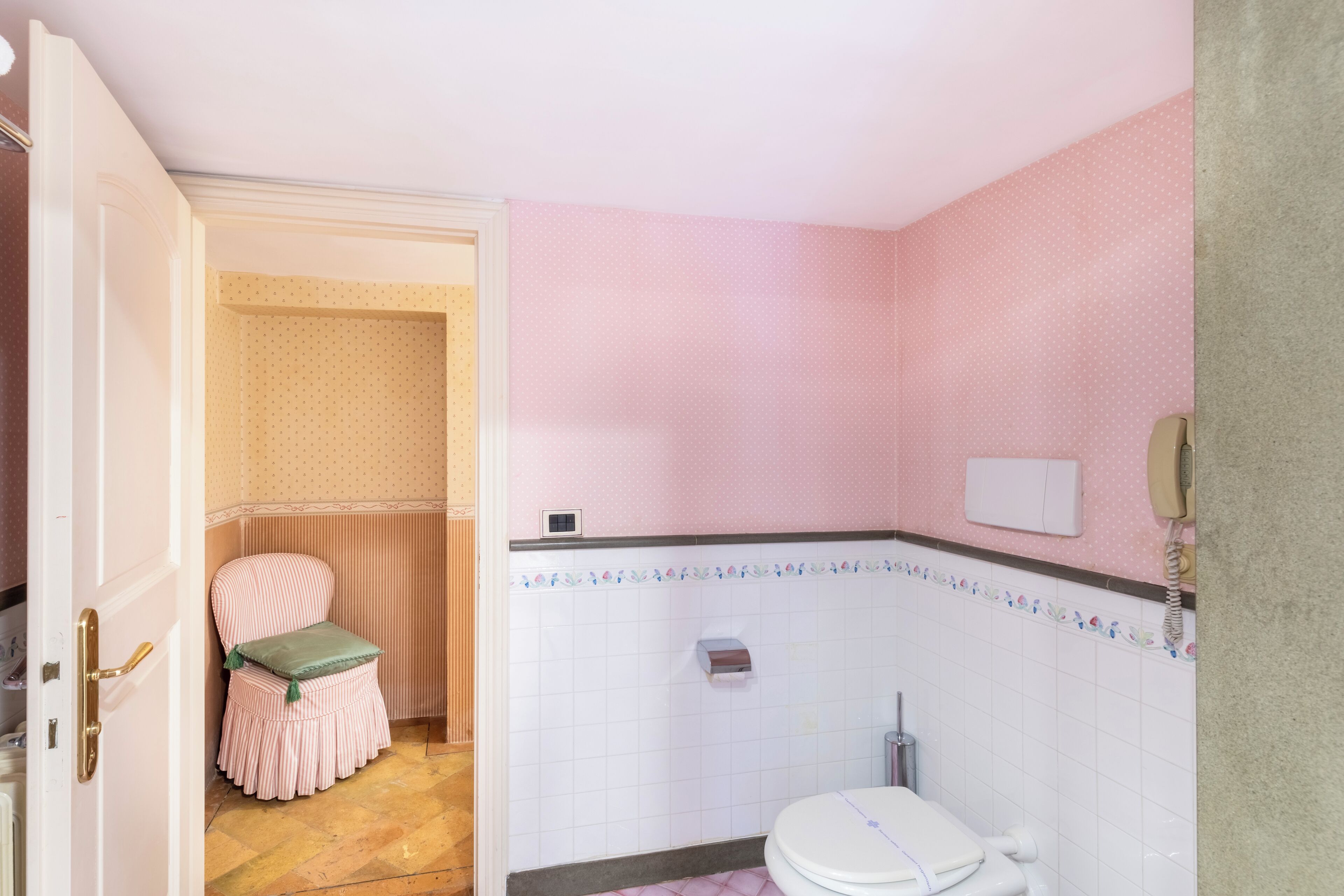 Een badkamer met een toilet, wastafel en douche. Er staat een stoel met een groene handdoek en een gestreept kussen. De muren zijn roze met een stippenpatroon.